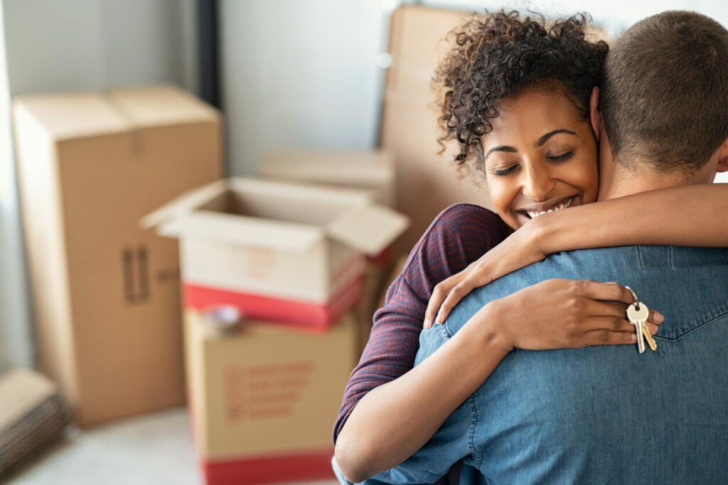 Woman hugging someone as they hold house keys with moving boxes in the background.