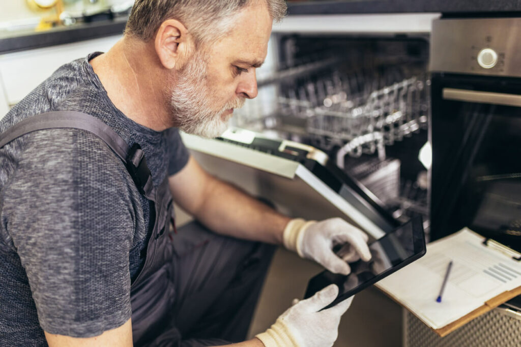 Man checking the inside of a dishwasher and marking tasks on a clipboard.