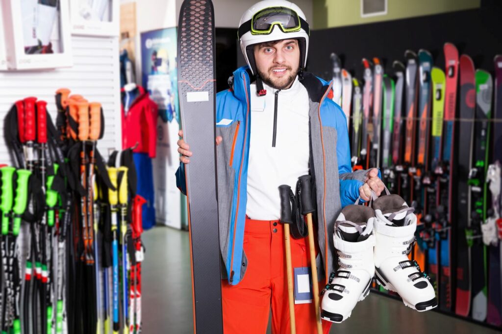 A smiling man in skiing gear inside of a sports equipment shop.