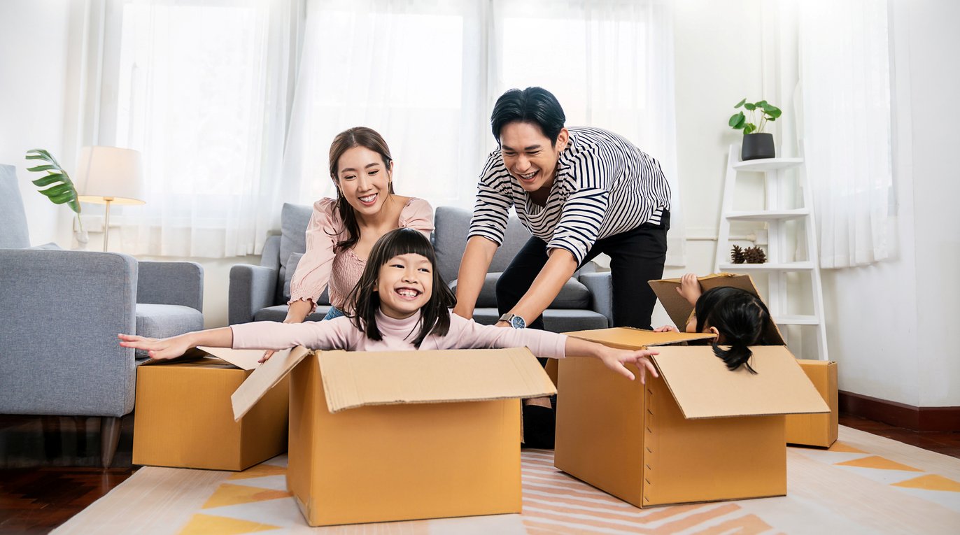 A happy family playing with moving boxes in a bright living room with white walls, sheer white curtains, and gray couches.