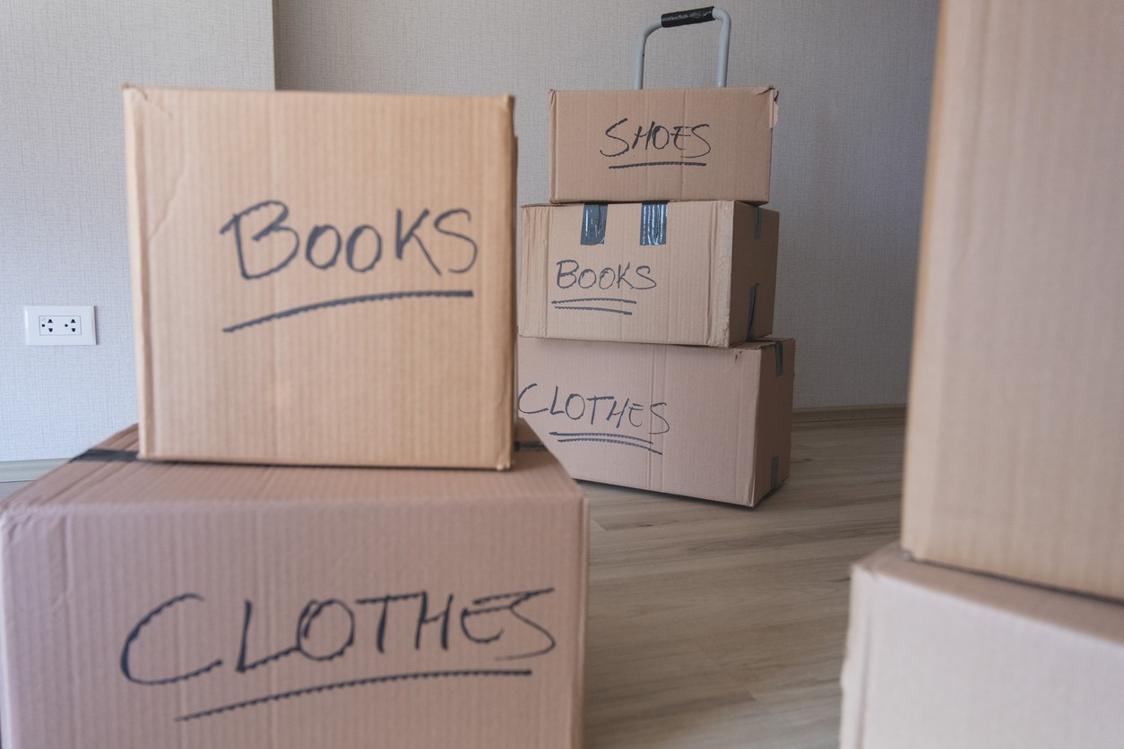 Stacked moving boxes labeled for books, clothes, and shoes inside a new home, on light wood flooring.